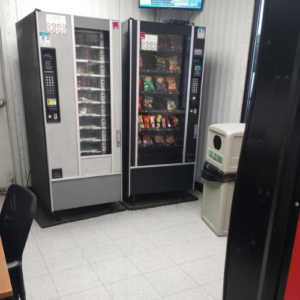 Two vending machines stand in a room beside a table, offering snacks and drinks for visitors.
